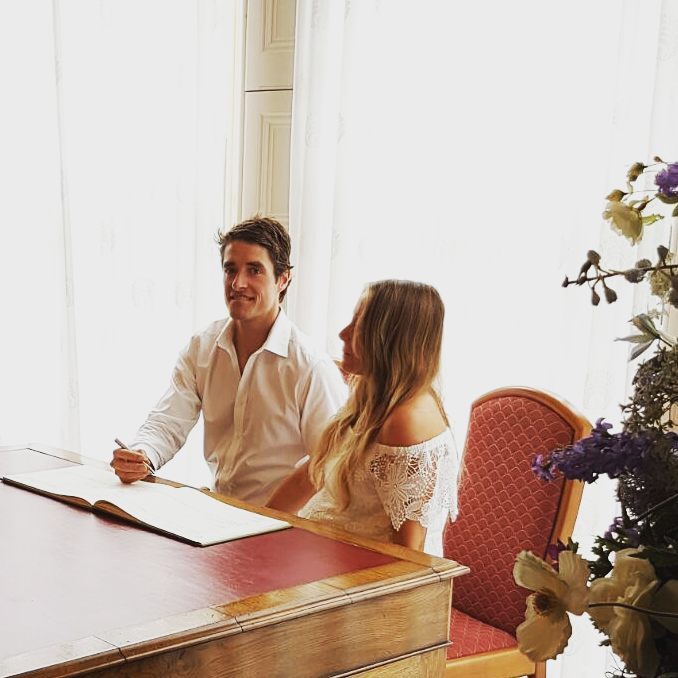Man signing a document at a wooden desk beside a woman in white lace, seated by a window with flowers nearby.