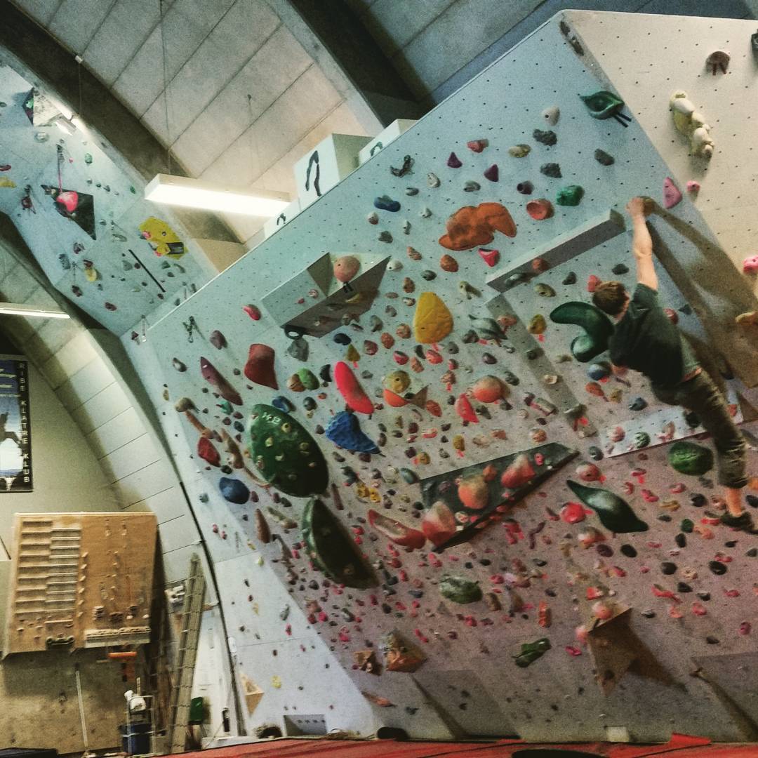 Indoor climbing gym with an overhanging wall covered in colorful holds; a climber ascends near the top right.