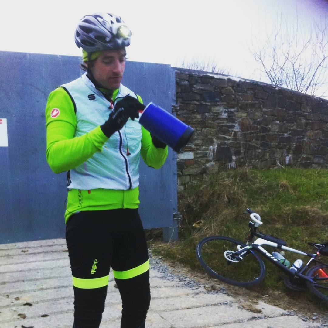 Cyclist in helmet and neon jacket holding a blue container, standing near a bicycle lying on grass beside a stone wall.