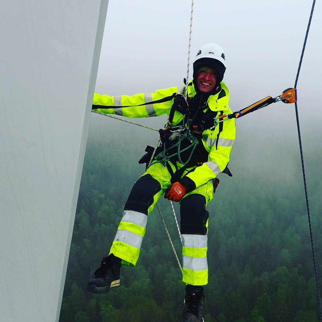 Rope-access technician in high-visibility suit and helmet suspended by ropes beside a wind turbine blade, forest visible below in mist.