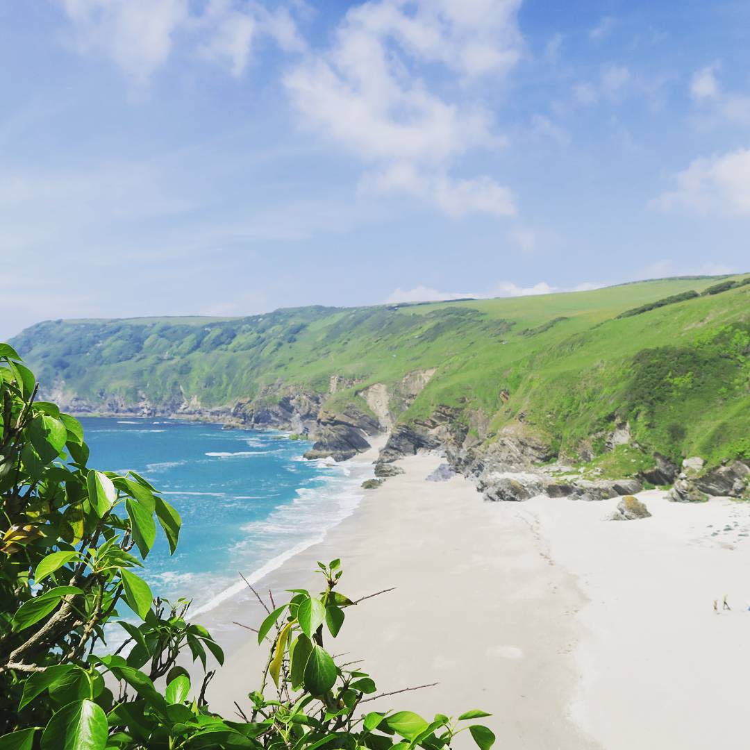 Coastal view of a sandy beach and turquoise sea bordered by green cliffs, with foliage framing the foreground and a partly cloudy sky above.