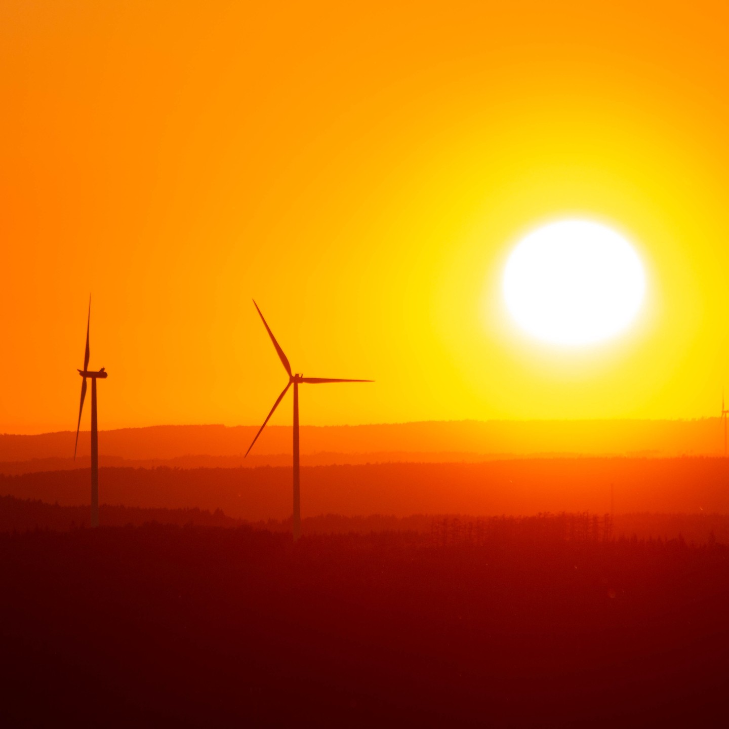 Silhouettes of two wind turbines against an orange sky with a bright sun low near the horizon, dark hills below.