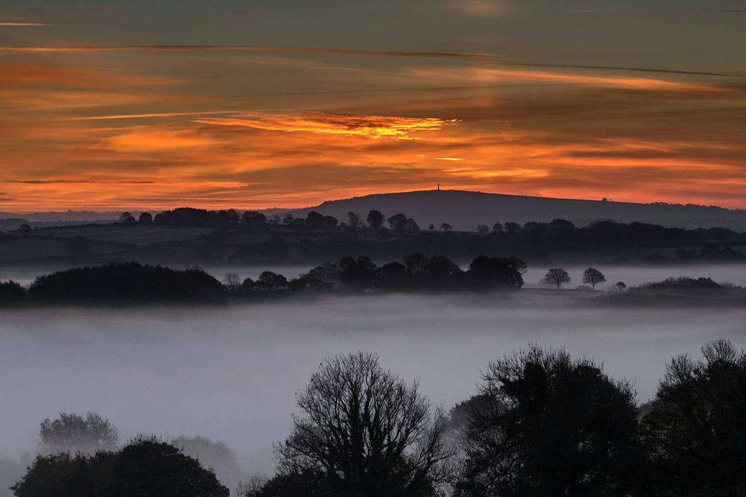 Layer of ground mist over fields at dawn, silhouetted trees and distant hill with monument, orange sunrise sky above.
