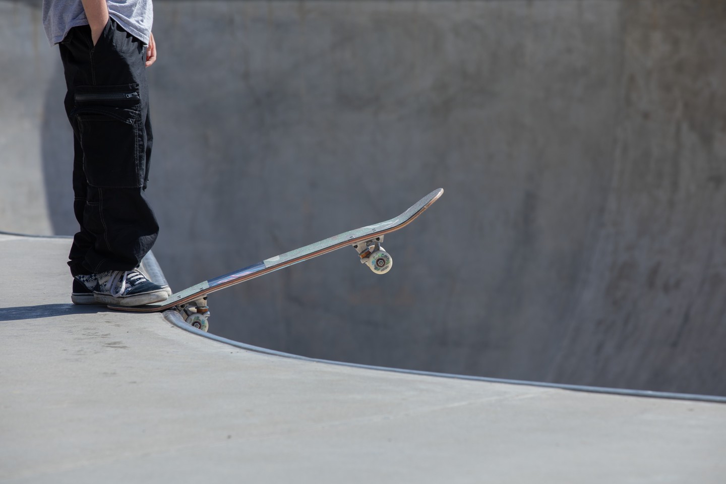 Skateboarder standing at edge of concrete skate bowl, skateboard tipped on its tail at the lip, one wheel airborne.