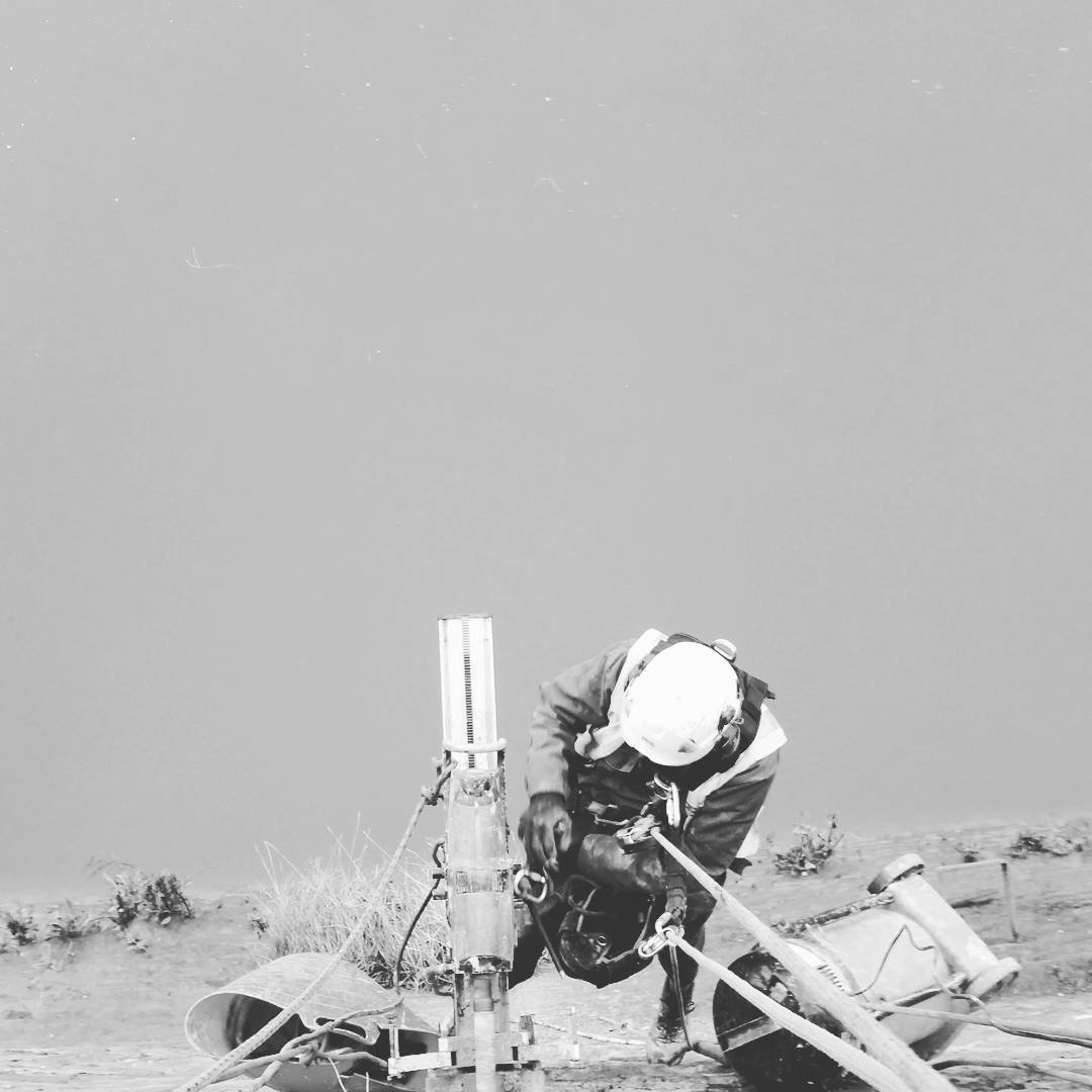 Worker in helmet and harness bends over equipment on rope, near drilling rig and pipes, misty outdoor site, black and white.