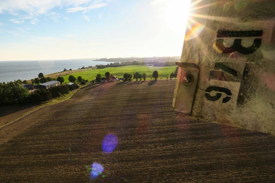 Aerial view from a wind turbine tower showing farmland, coastline, and trees, with metal panel and lettering in foreground.
