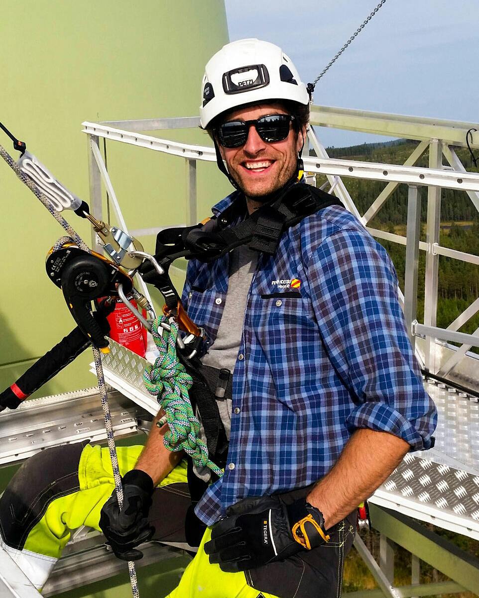 Smiling man in helmet, sunglasses, and plaid shirt wearing climbing harness with rope and gear, standing on wind turbine platform.