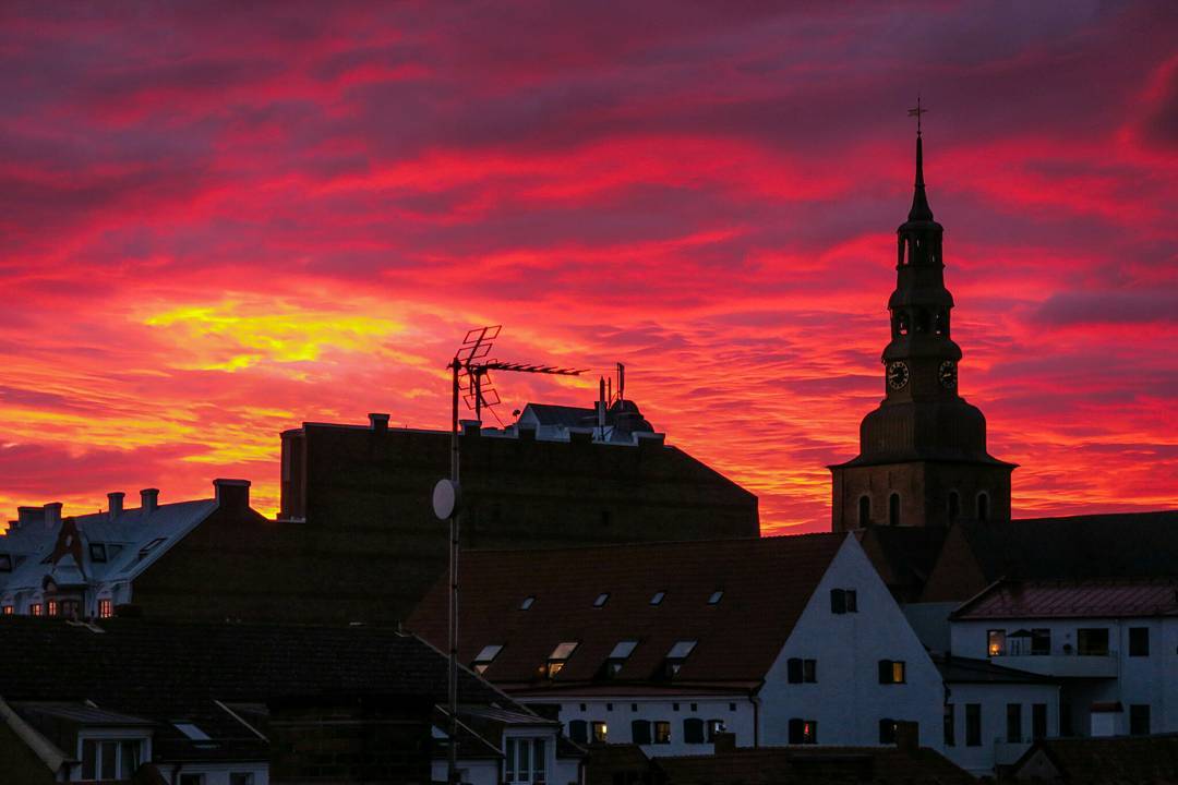 Silhouetted rooftops, a church clock tower, and a TV antenna against a vivid pink and orange sunset sky in Ystad, Sweden.