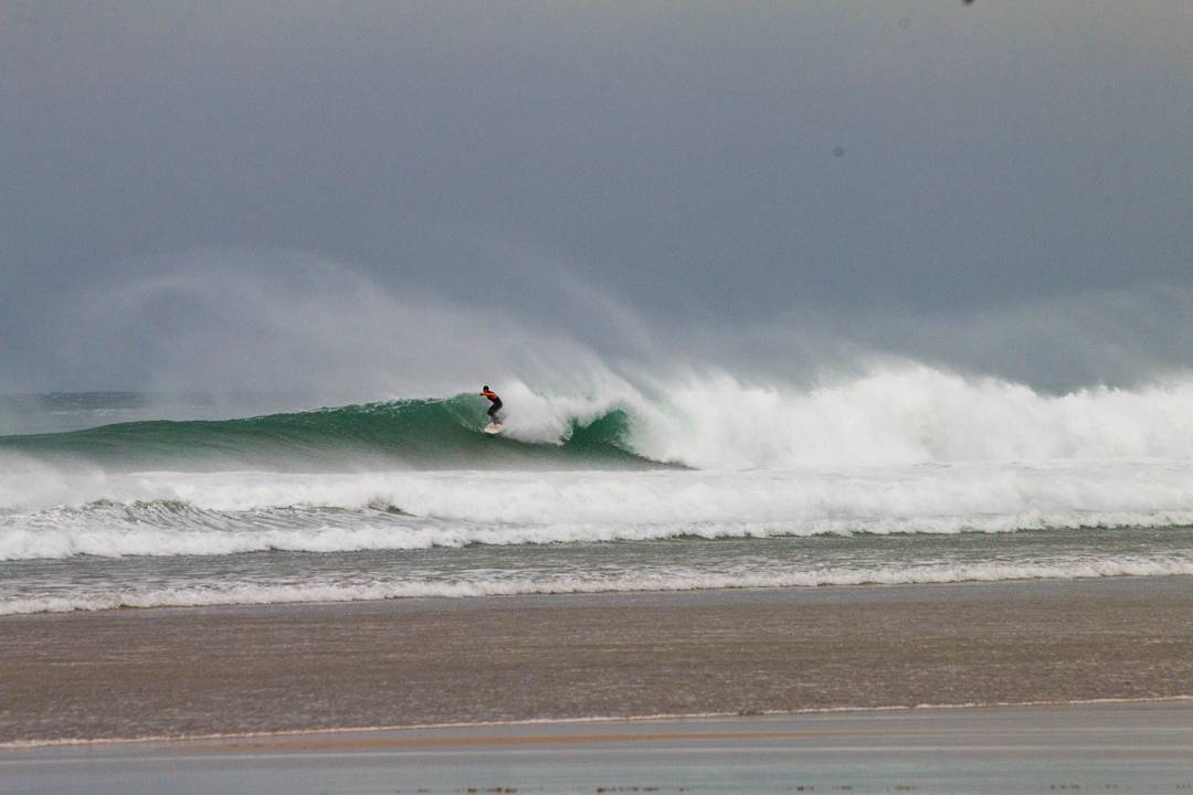 Surfer riding a large breaking wave, spray blowing off the crest, wide sandy beach in foreground, overcast sky.