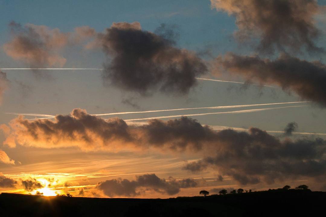 Sunrise sky with orange glow near horizon, scattered clouds, jet contrails crossing the sky, and a dark tree-lined hillside silhouette below.