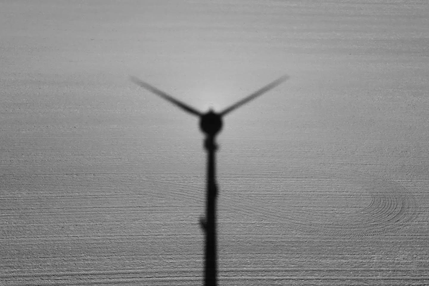 Black and white photo of a wind turbine silhouetted against the sky, viewed from below, with a plowed field in the background