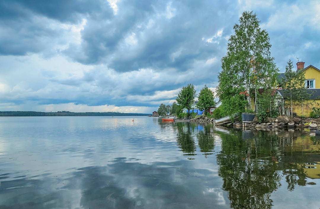 Calm lake shoreline with yellow house, trees, and a small boat under a cloudy sky, reflections visible in still water.