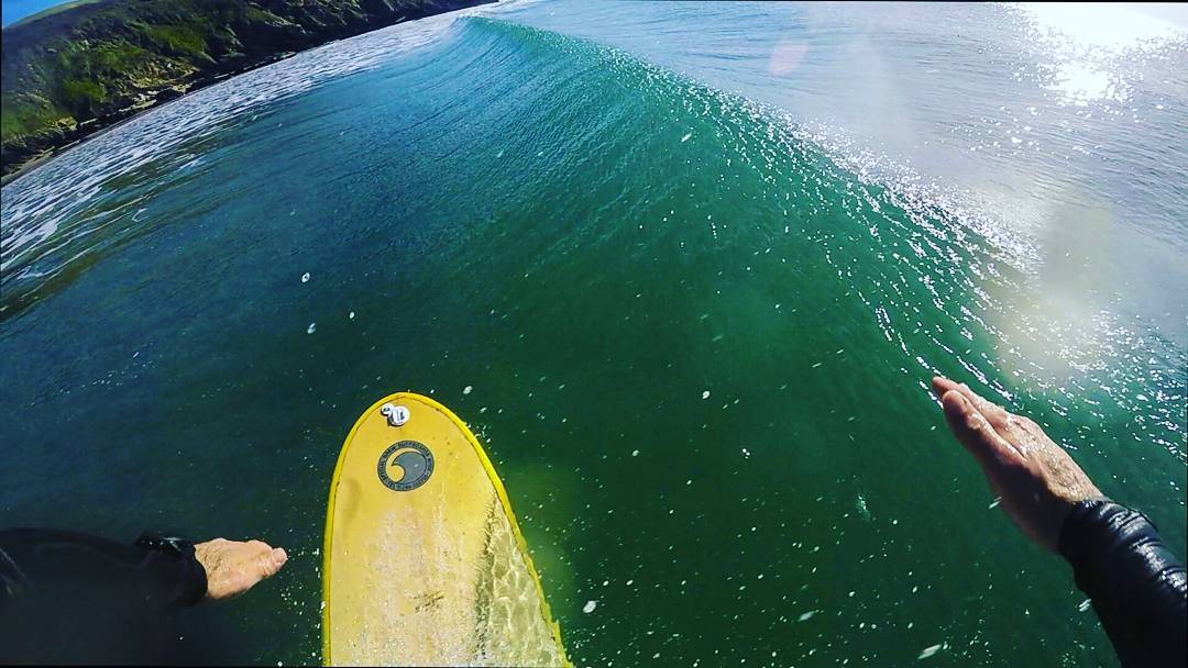 POV shot from a surfboard on green ocean water, paddler's hands visible, breaking wave and coastline in background.