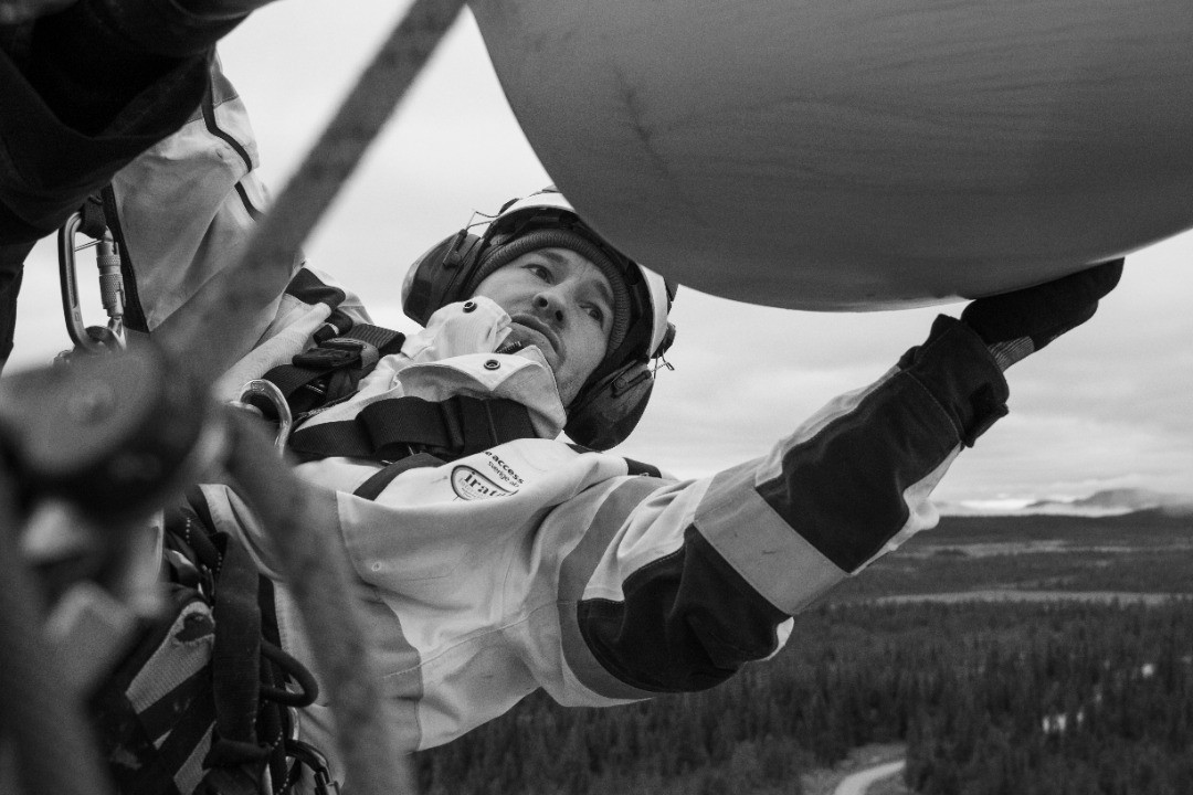 Black-and-white photo of a rope-access technician in safety harness inspecting a wind turbine blade high above a forest.