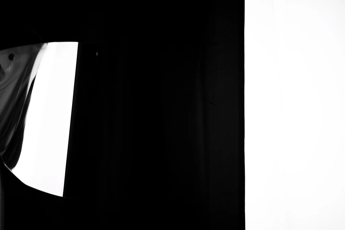 High-contrast black and white image with a dark curtain on the left, a bright white panel on the right, and a reflective curved surface catching light on the far left edge.