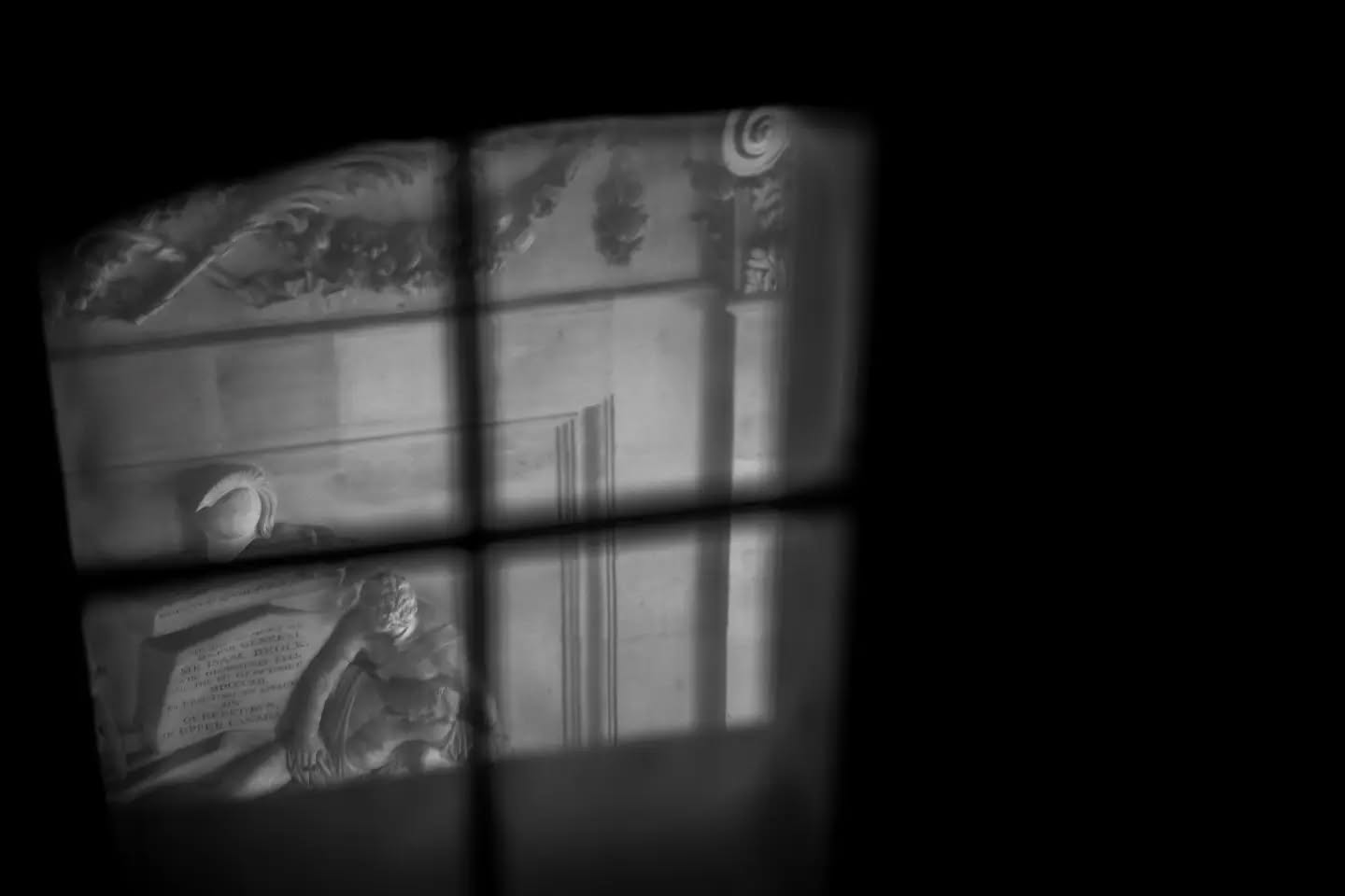 Black and white photo of a classical stone monument with reclining figure and carved inscription, seen through a window with dark cross-shaped frame.