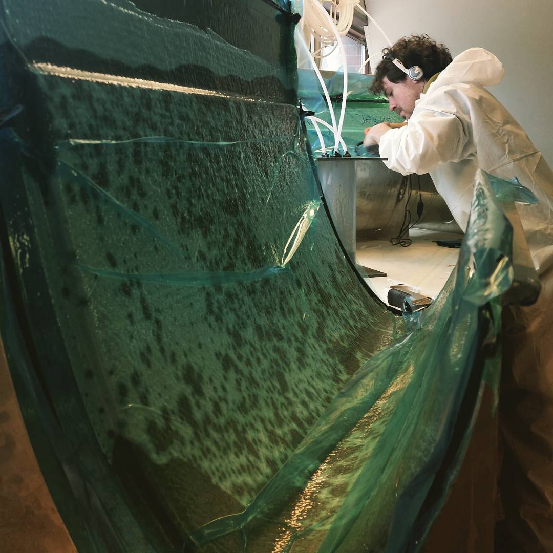 Person in white protective suit and headphones working on large curved sheet of textured green glass indoors.