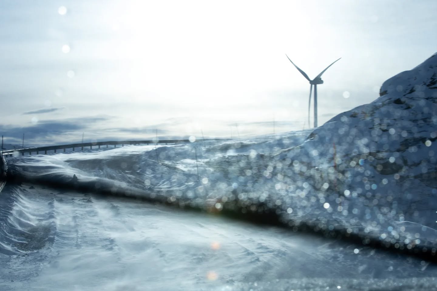 Snow-covered road with guardrail leading to a wind turbine, viewed through a frosted, water-droplet-covered window.