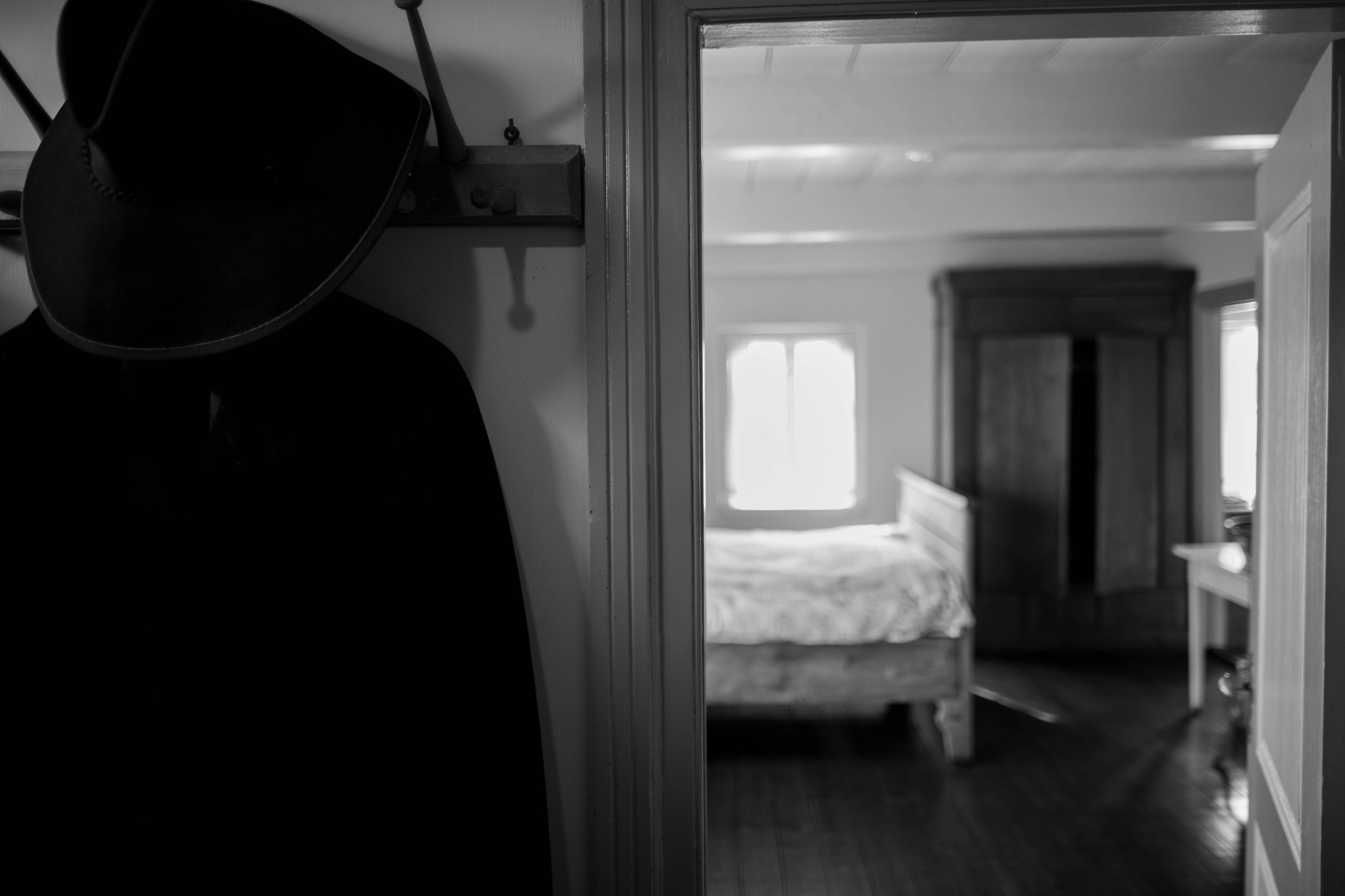Black and white photo of a dark coat and hat on wall hooks beside a doorway leading to a sunlit bedroom with bed and wardrobe.