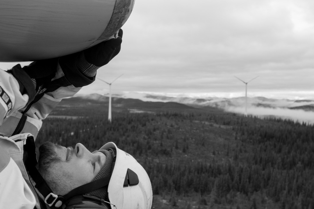 Black-and-white photo of a helmeted worker looking up at a wind turbine blade, with forested hills and turbines below.