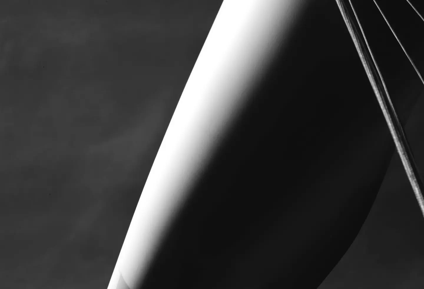 Black and white close-up of a curved surface with a bright diagonal highlight, thin cables visible in the background on the right.
