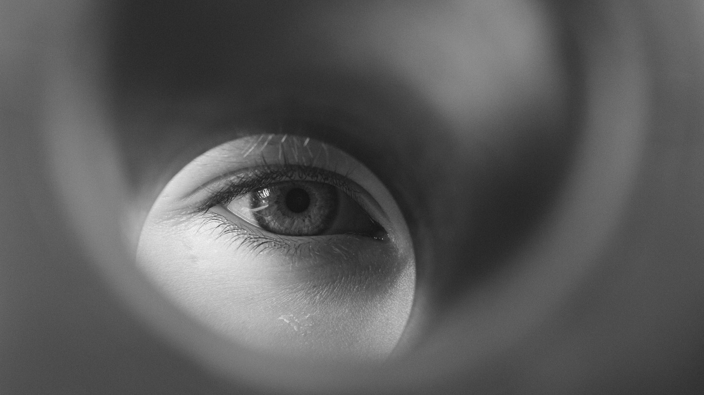 Black-and-white close-up of a human eye viewed through a circular tube or cylindrical opening, eyelashes visible.