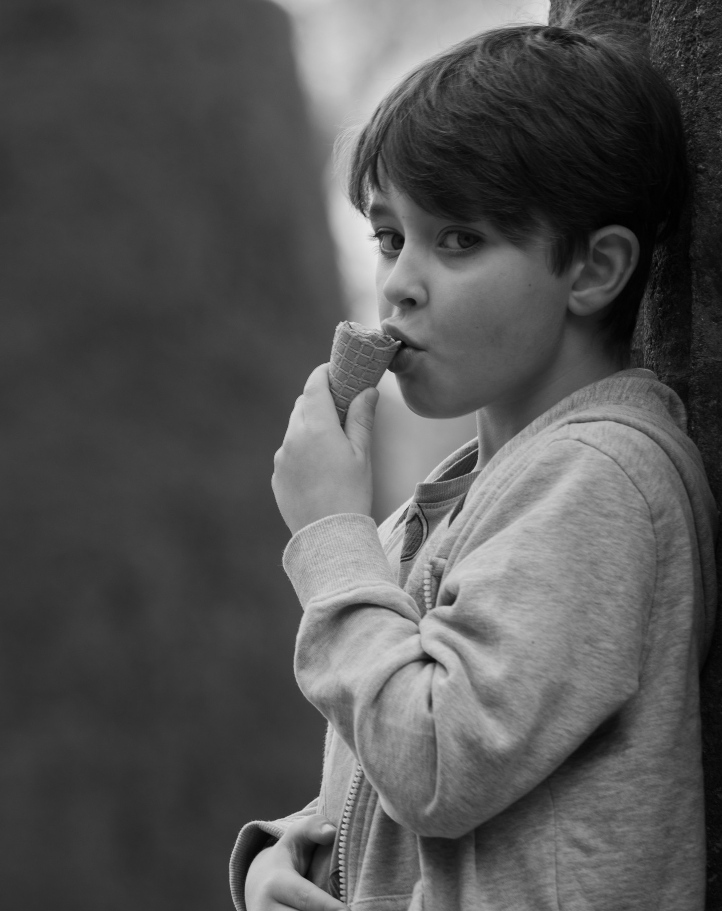 Black-and-white portrait of a boy in a hooded sweatshirt eating an ice cream cone, looking sideways at the camera.