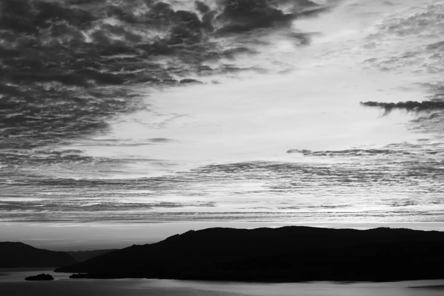 Black and white photo of layered clouds over a calm lake, with dark silhouetted hills and small islands in the foreground.