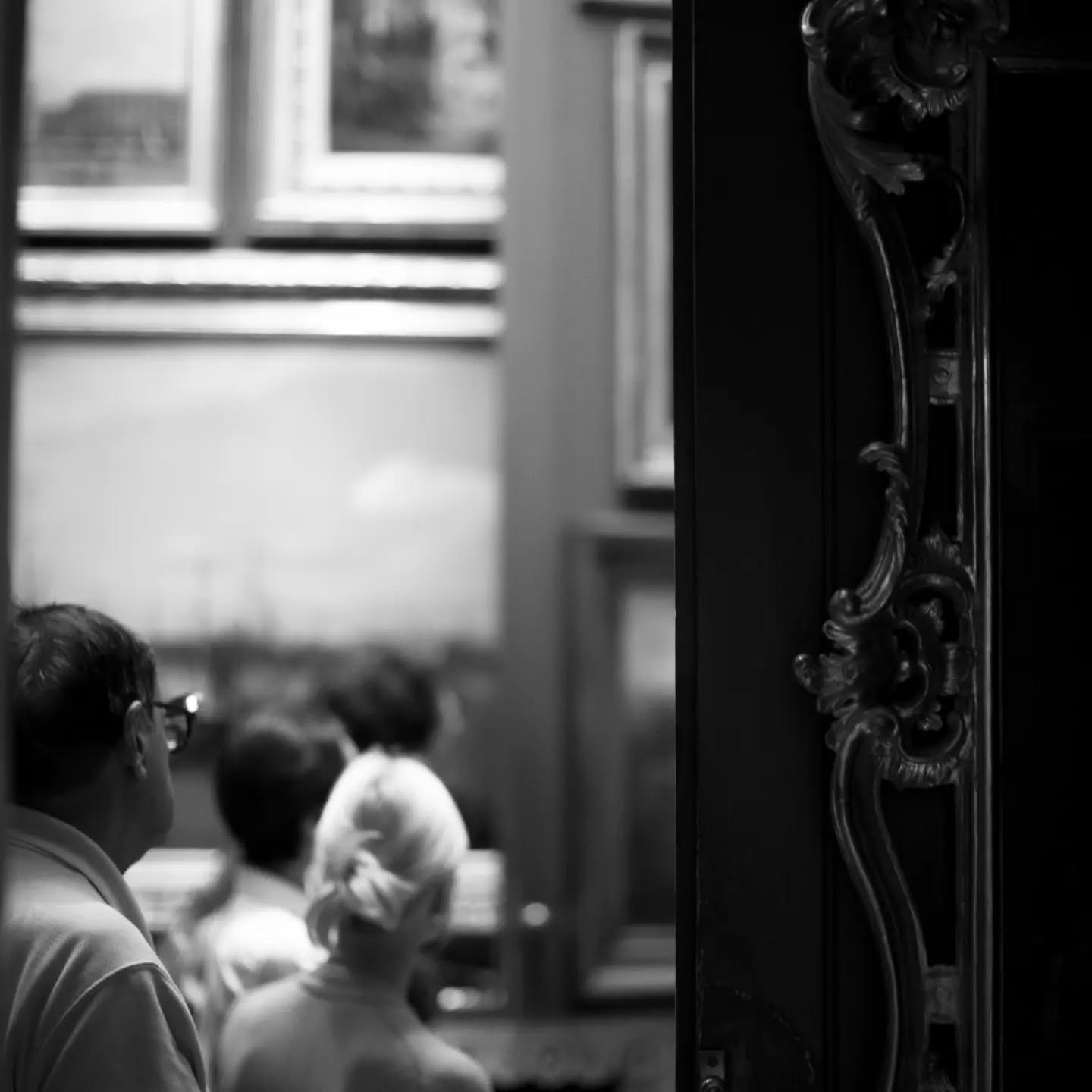 Black-and-white photo of people viewed from behind near a window, with an ornately carved dark wood column in the foreground.