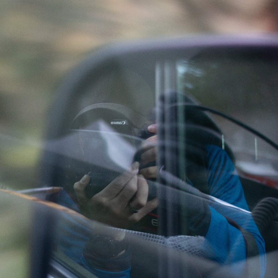 Reflection in a car side mirror showing a person holding a Canon camera up to their face, wearing a blue shirt.