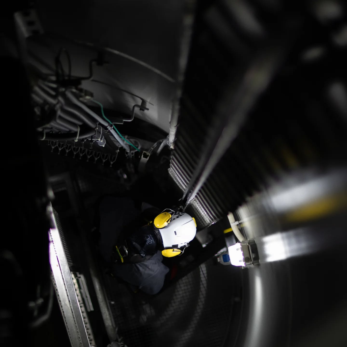 Worker in white helmet with yellow ear protection inspecting inside a narrow industrial machine or turbine, illuminated by a bright light.