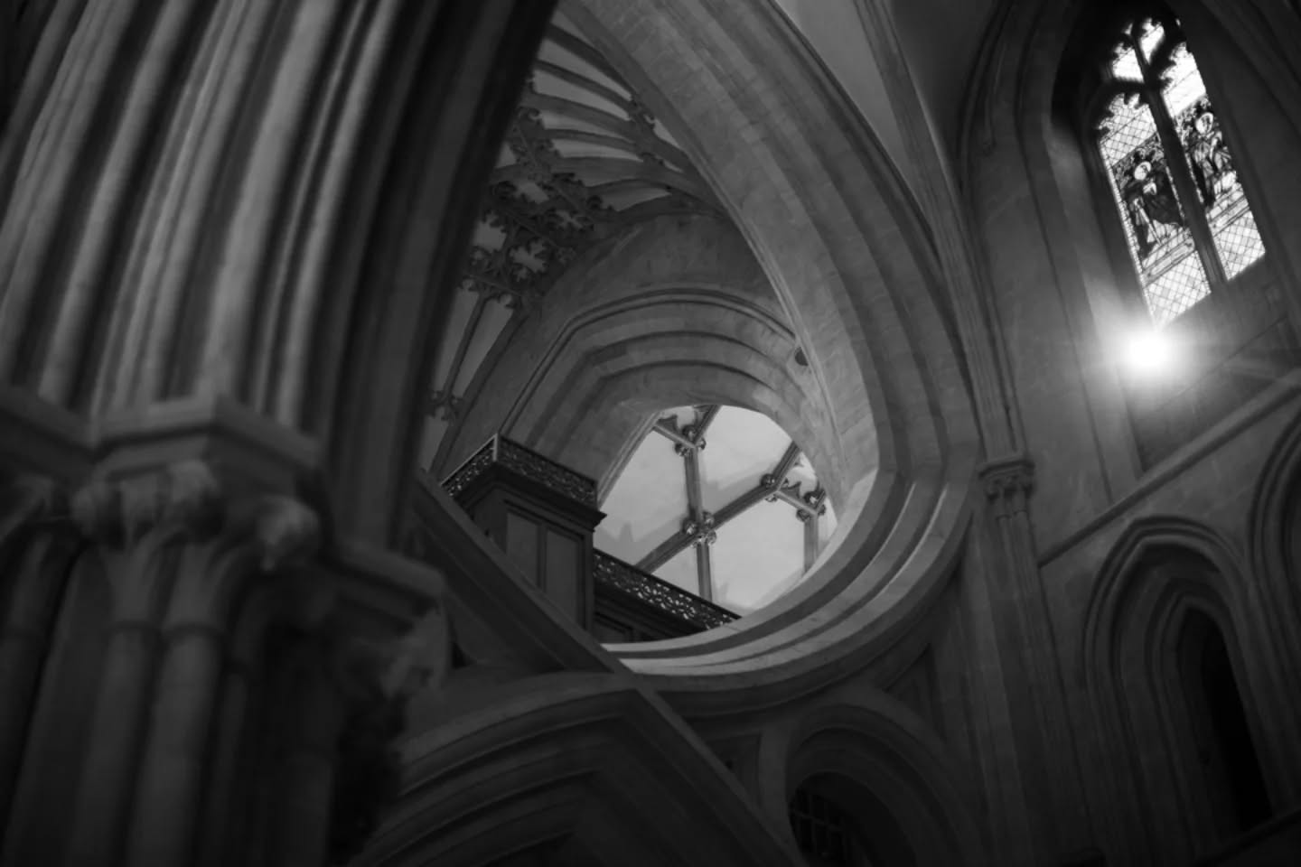 Black and white photo of Gothic cathedral interior, looking up at crossed stone arches, with stained glass window and sunlight at right.