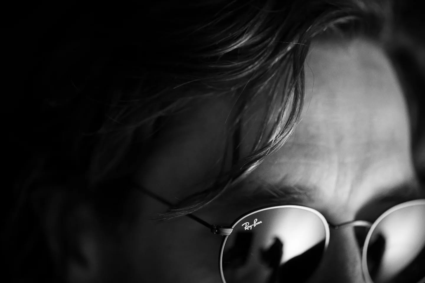 Black-and-white close-up of a person's forehead and windblown hair, wearing round Ray-Ban sunglasses reflecting light.
