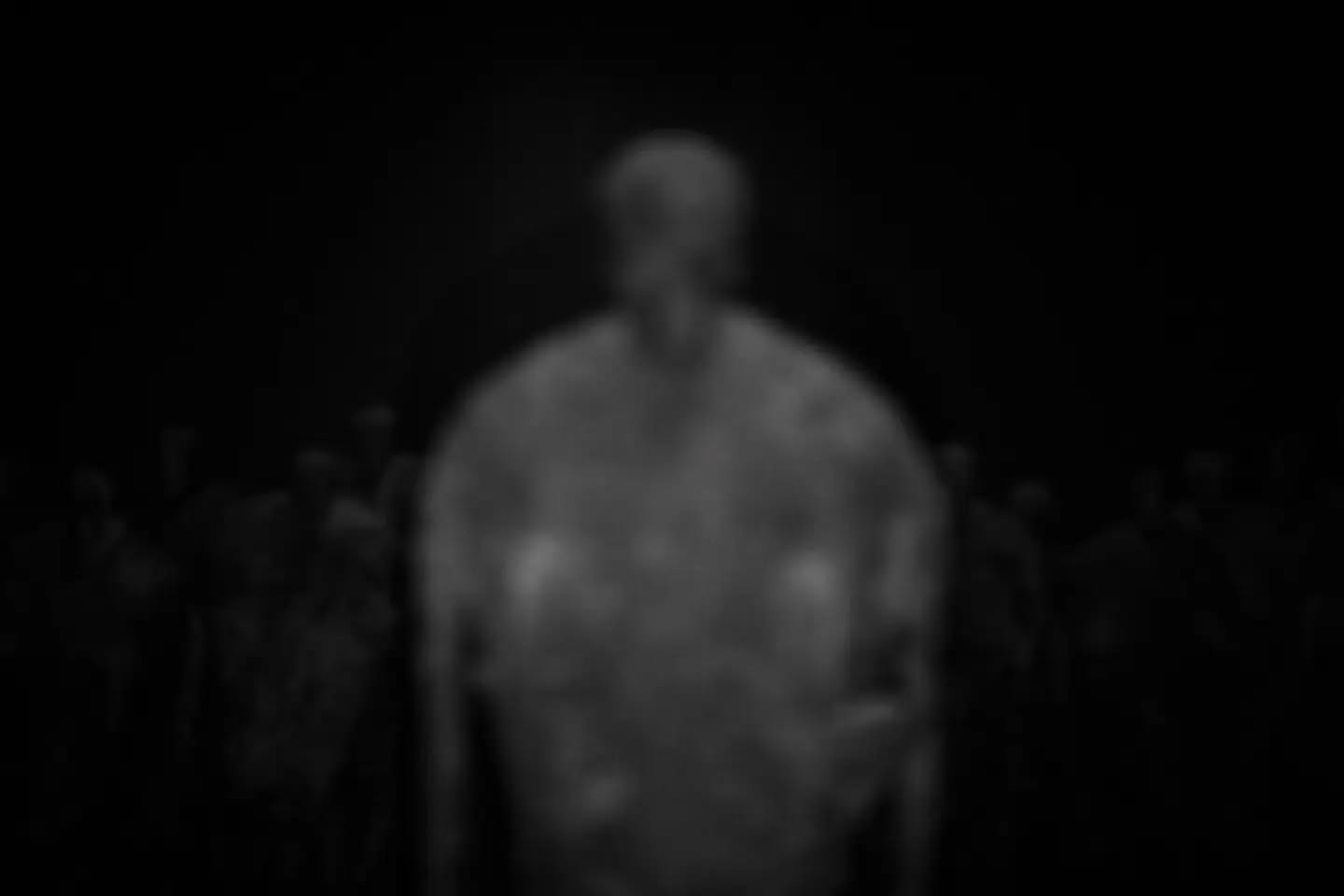 Blurred black-and-white photo of a shirtless figure facing forward, surrounded by faint silhouetted figures in the dark backg