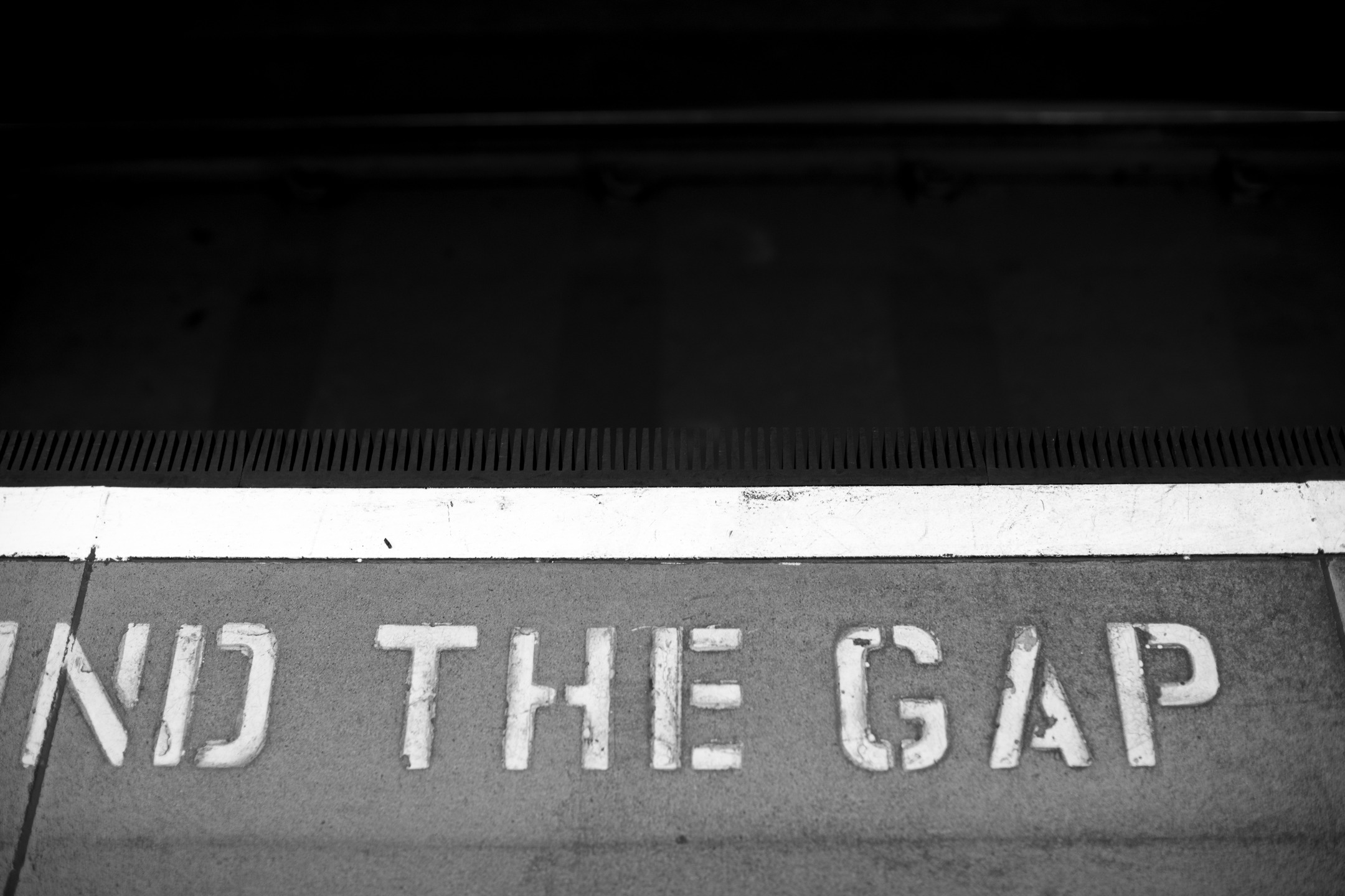 Black-and-white photo of "MIND THE GAP" stenciled on a train platform edge, with a white safety line and dark tunnel above.