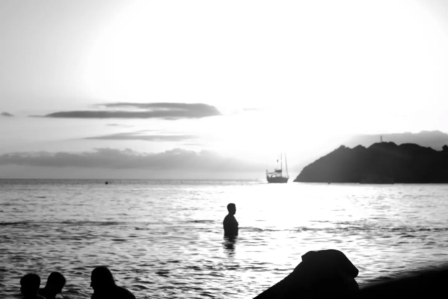 Black and white photo of a person standing waist-deep in the sea at sunset, with a sailboat and headland in the distance and 