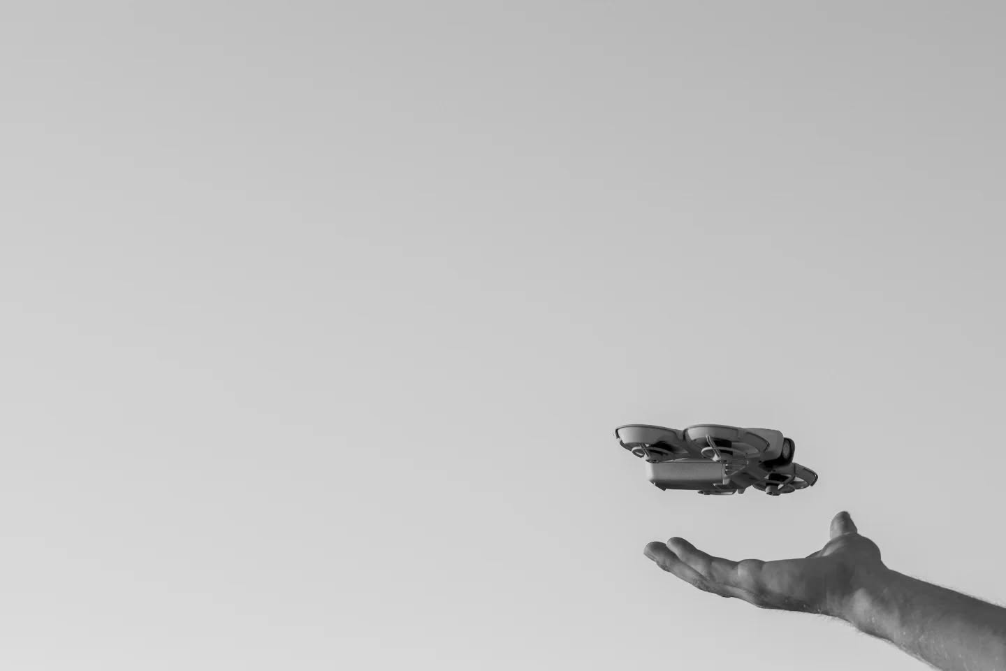 Black-and-white photo of a small drone hovering above an outstretched open hand against a plain gray sky.