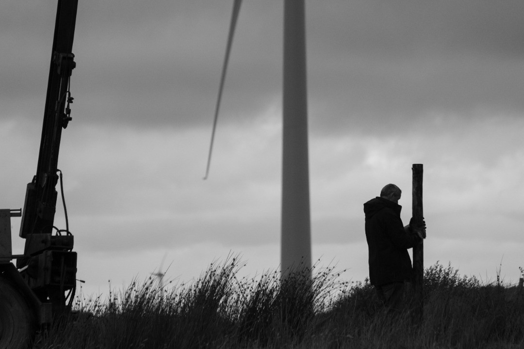 Black-and-white photo of a man holding a wooden post beside tall grass, with a wind turbine and crane in background.