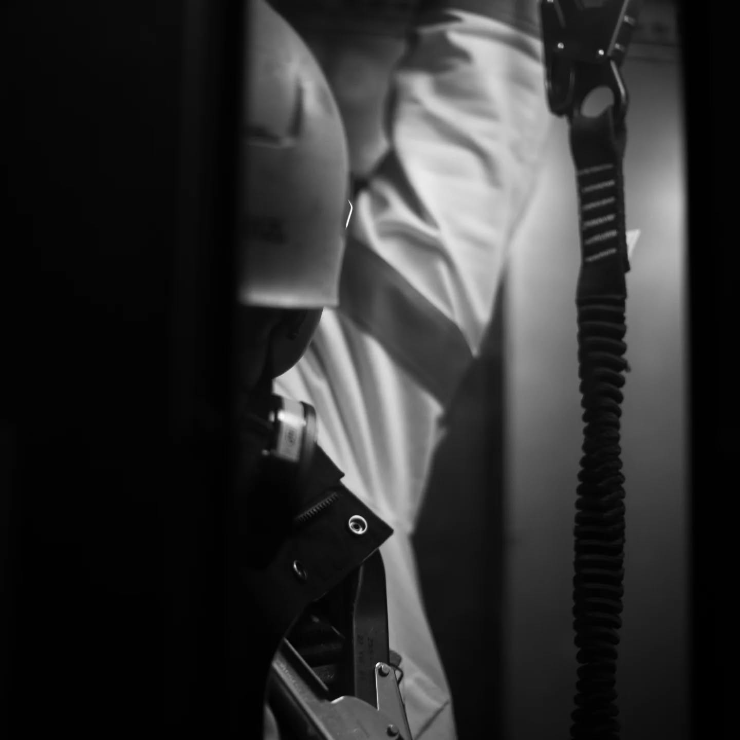 Black and white close-up of a hard hat, safety harness clips, and a coiled lanyard hanging in narrow, dim space.
