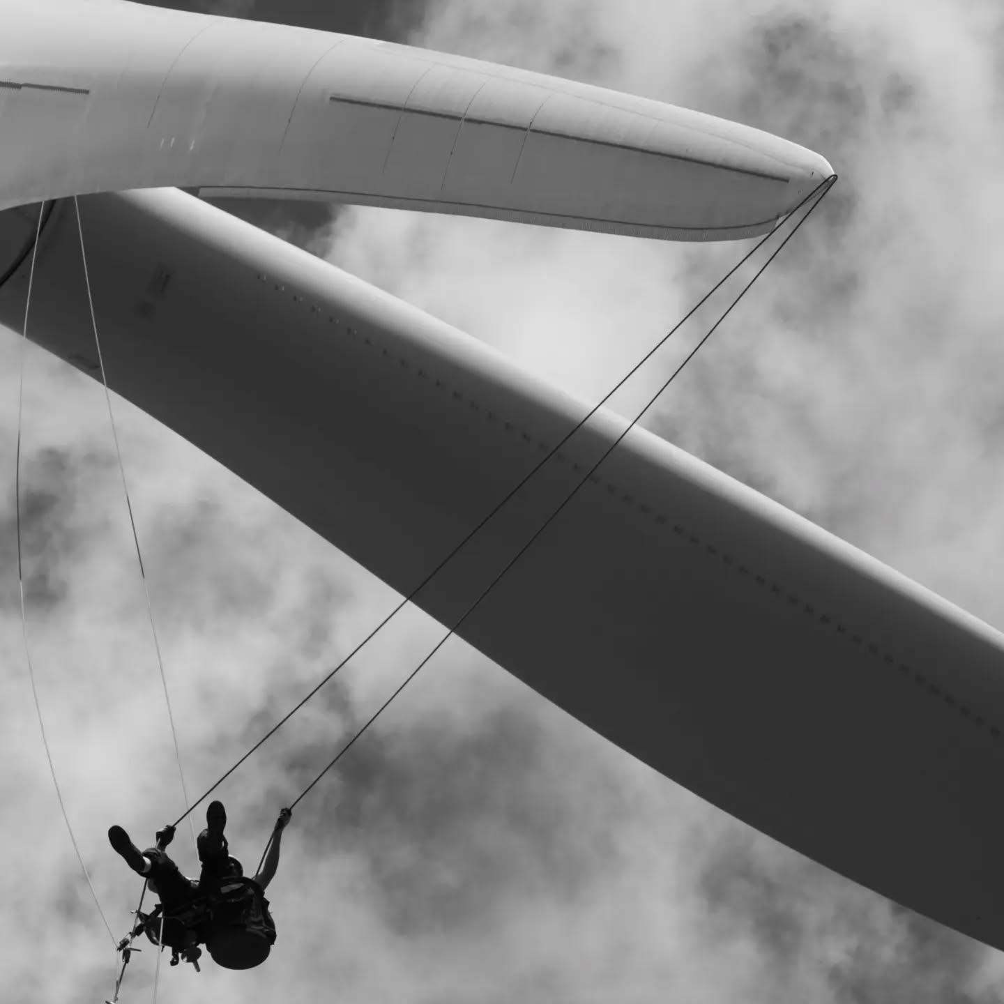 Black-and-white photo of a paraglider wing seen from below, with a silhouetted person hanging from the lines against a cloudy