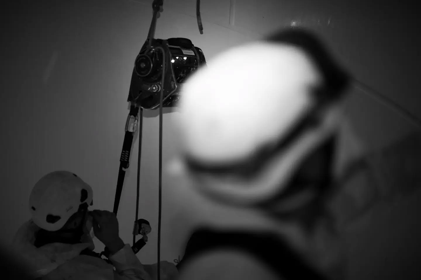 Black-and-white photo of a person in a helmet using rope and mechanical rigging equipment, with a blurred helmeted figure in 