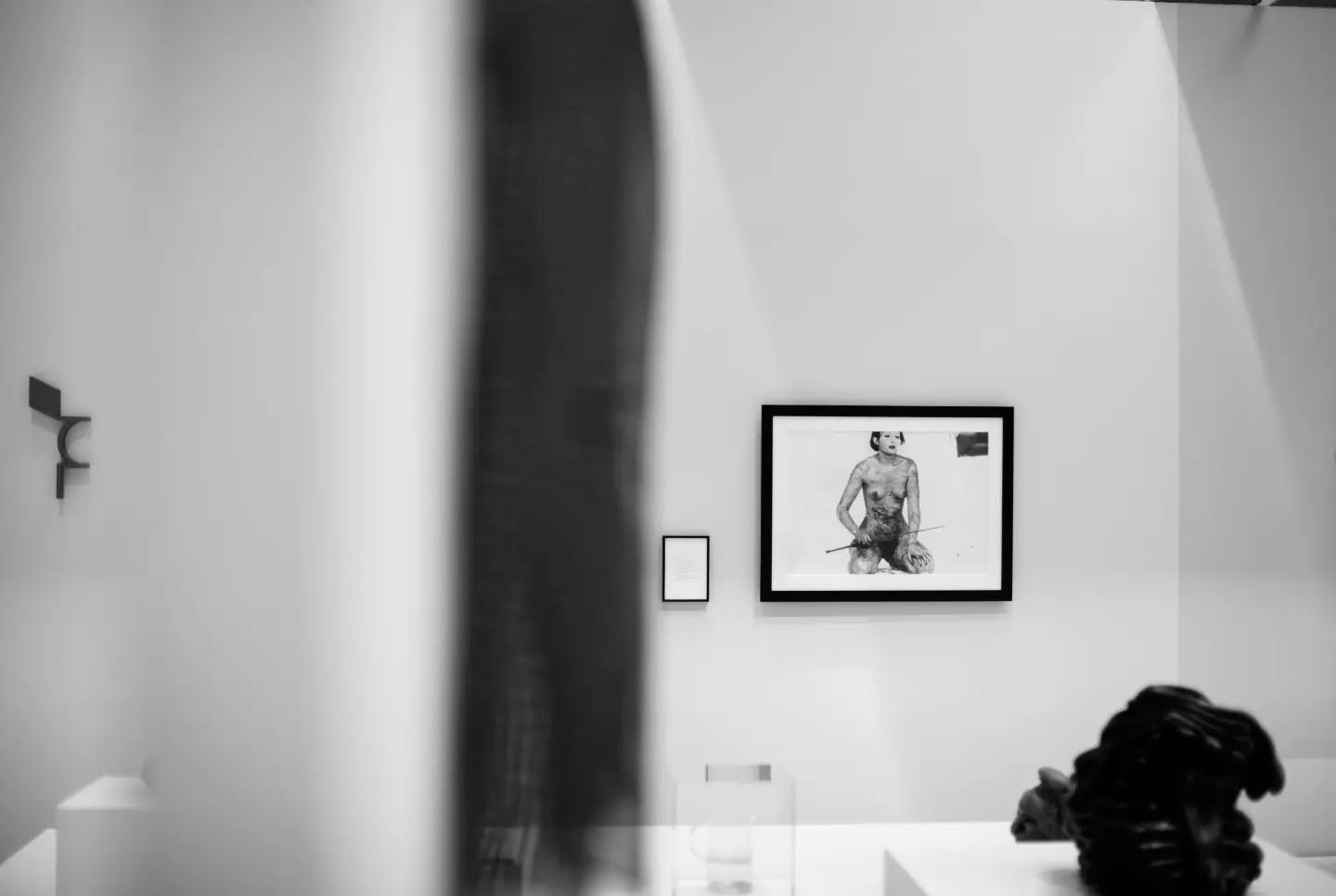 Gallery interior in black and white; a blurred figure in foreground, framed nude portrait and small plaque on wall, dark sculpture on table.