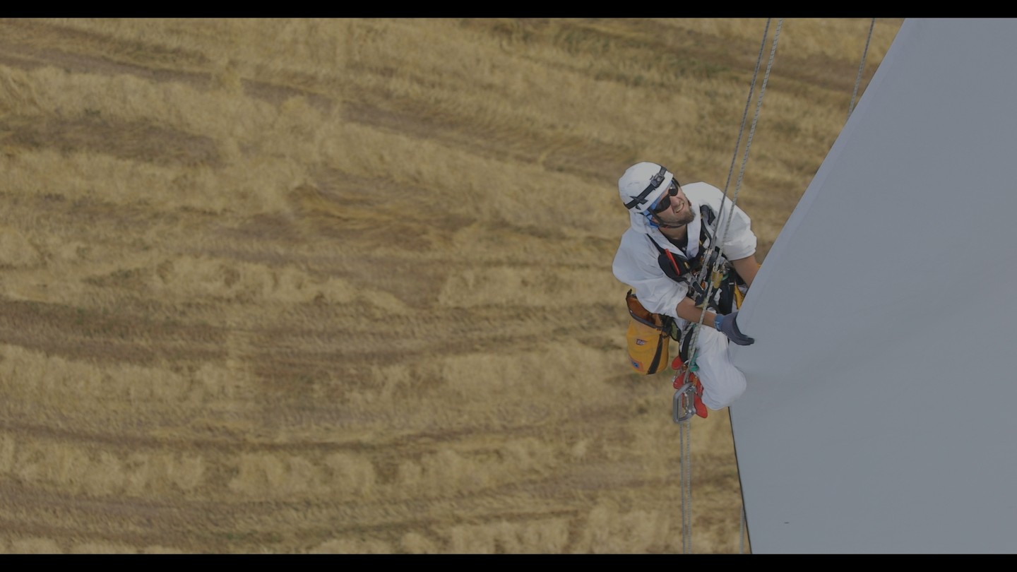Rope-access technician in white coveralls and helmet rappelling down a wind turbine blade above a golden field.