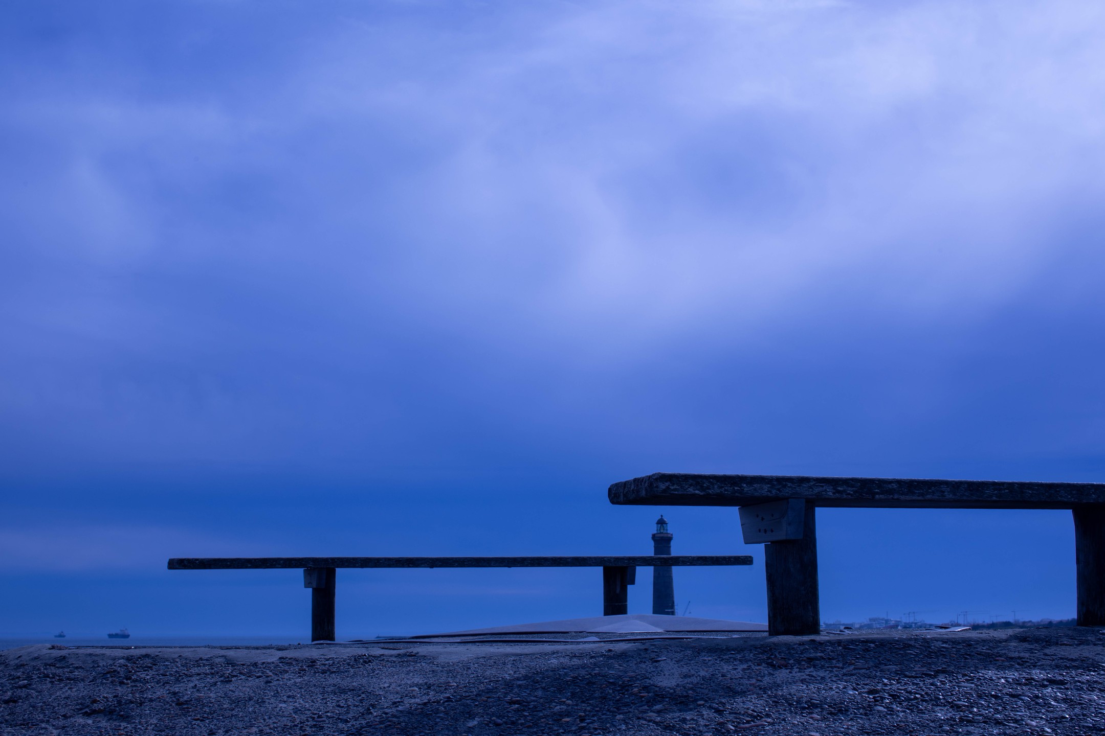 Two wooden benches on a gravel shore at dusk, with a distant lighthouse and ships under a cloudy blue sky.