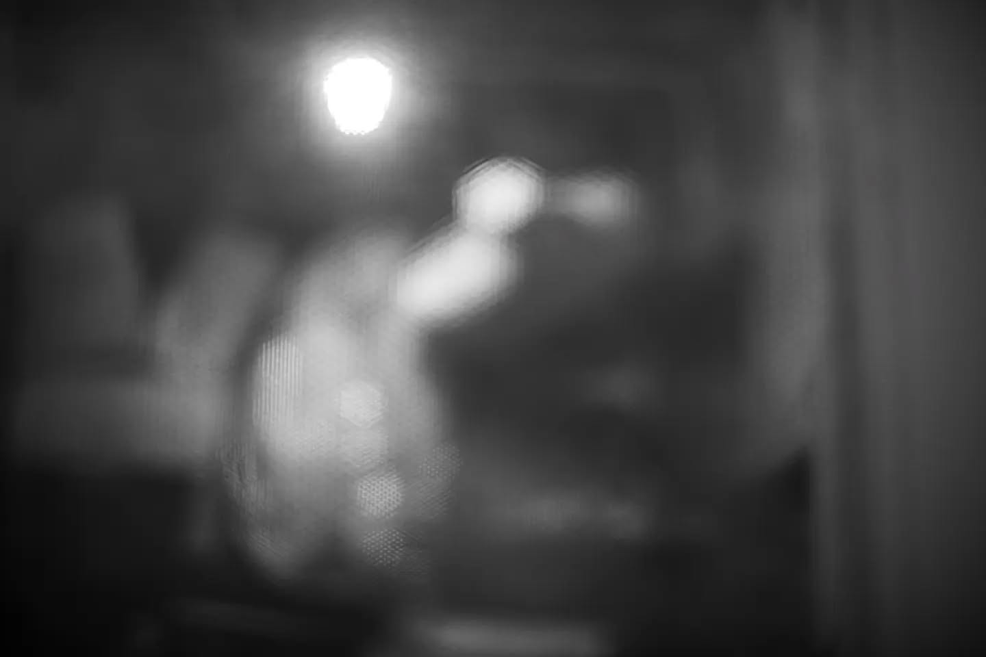 Blurred black-and-white photo of a figure viewed through a textured screen, with a bright light source overhead and an extend