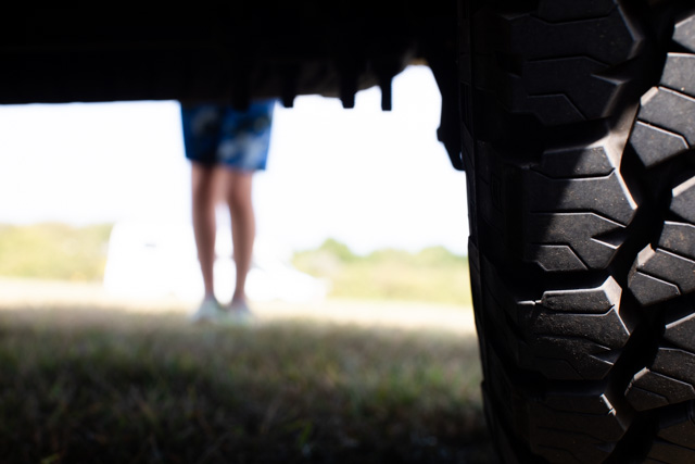 View under a vehicle showing a large tire in focus, with a blurred figure in shorts standing in the sunlit background.