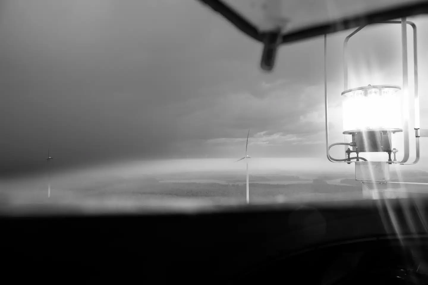 Black-and-white image of an illuminated aviation obstruction light on a structure, with a distant wind turbine visible over a hazy horizon.
