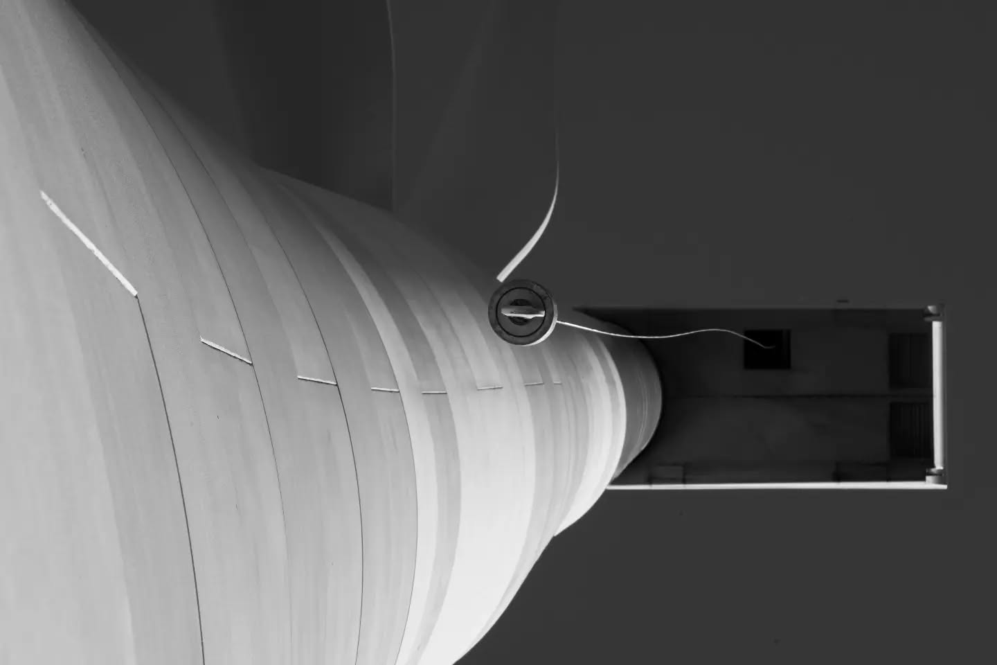Black-and-white close-up of a curved, ribbed cylindrical structure with a small round vent, extending into a rectangular metal duct or corridor.