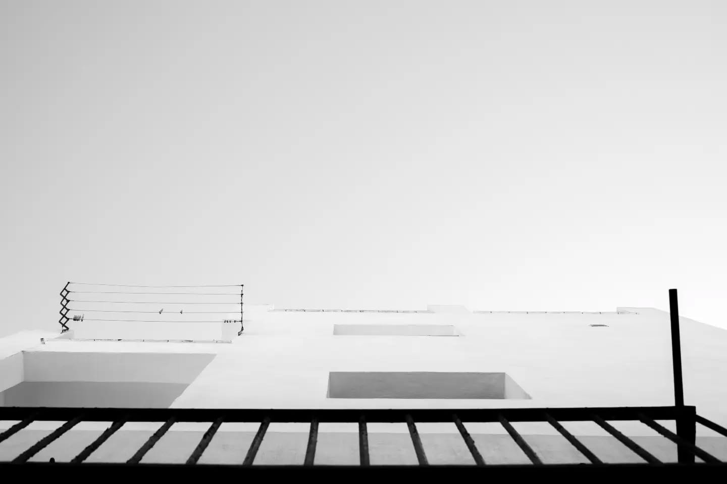 Black-and-white photo of a white building facade with recessed windows, wire fencing above, seen behind a railing.