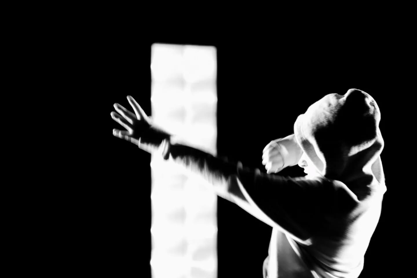 Black and white image of a hooded figure with arm extended, hand open, motion blur, against a bright vertical light source.